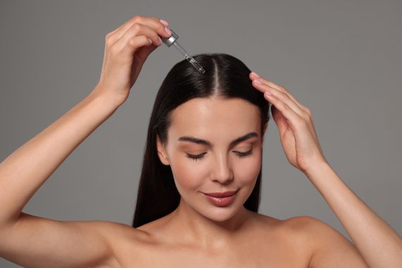 Mooie vrouw die haarserum aanbrengt op een grijze achtergrond. Cosmetisch product.